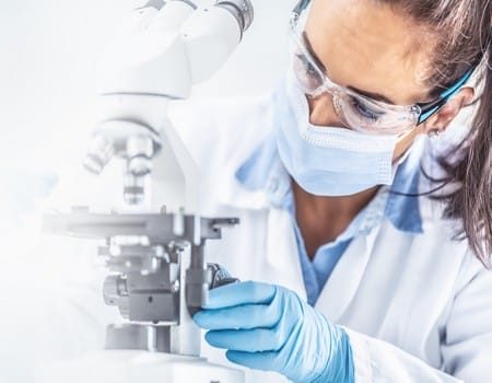 Female lab technician in protective glasses, gloves and face mask sits next to a microscope and conical flask, looking aside on the desk - panoramic banner.